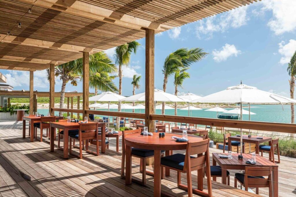 Um pátio de restaurante à beira-mar no Ambergris Cay Private Island com mesas e cadeiras de madeira, sombreado por um teto de ripas. Guarda-sóis brancos e palmeiras cercam a vista. Esse é um dos melhores resorts all inclusive no Caribe.