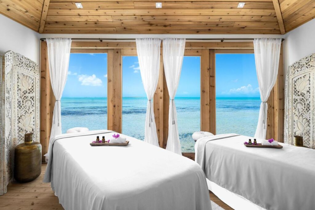 Duas mesas de massagem em uma sala do Ambergris Cay Private Island com cortinas transparentes e grandes janelas com vista para o oceano azul claro.