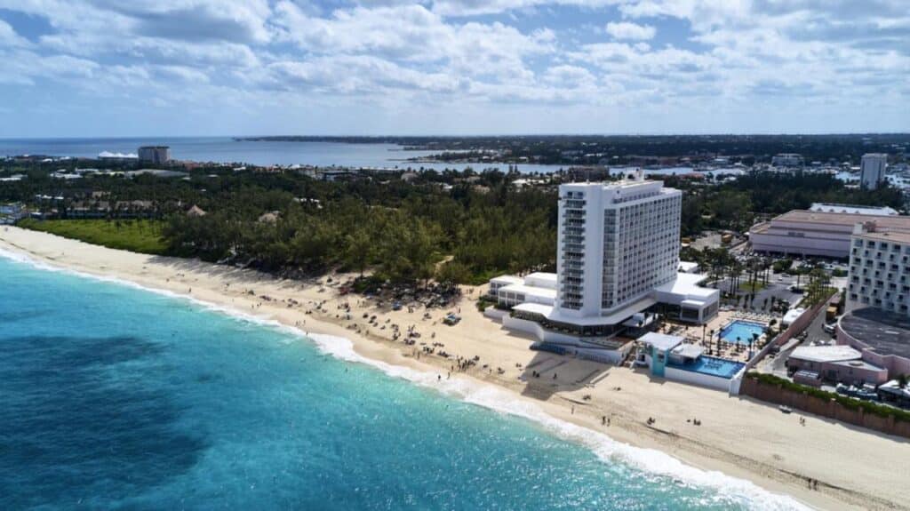 Vista aérea do Riu Palace Paradise Island à beira-mar, ao lado de uma praia de areia e oceano azul-turquesa, sob um céu parcialmente nublado com terra ao longe. Esse é um dos melhores resorts all inclusive no Caribe.