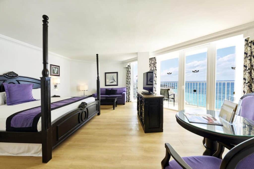 Quarto espaçoso do Riu Palace Paradise Island com cama de dossel, sofá e mesa de frente para uma varanda com vista para o oceano.