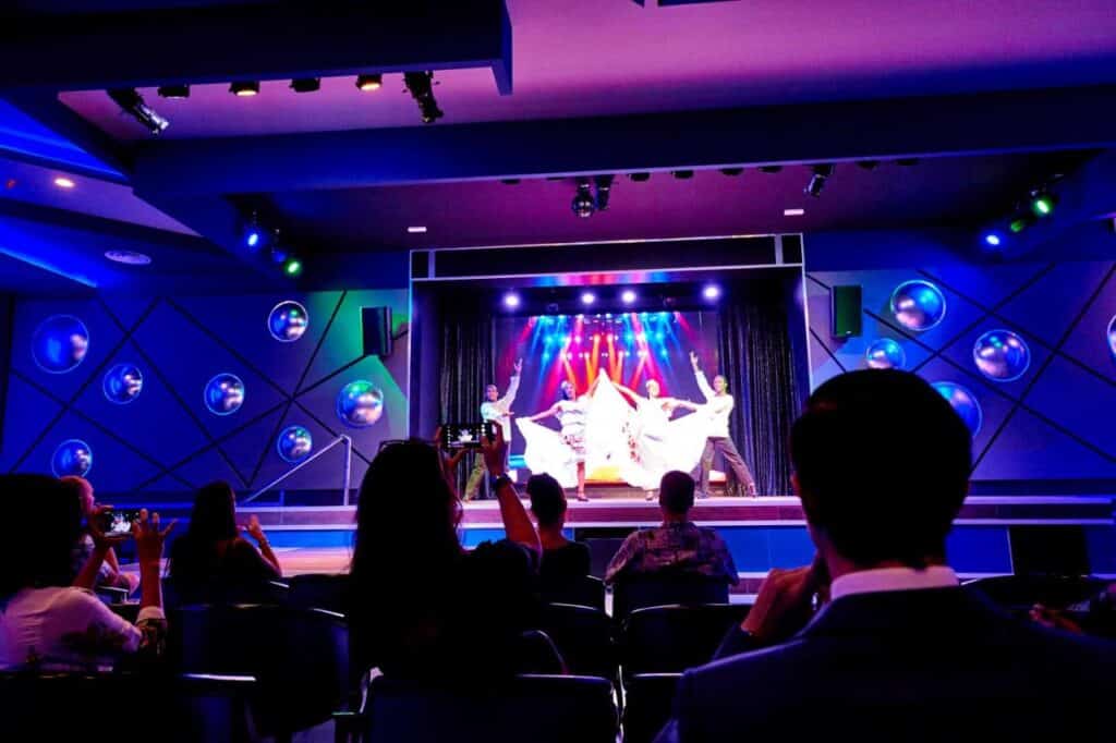 Um grupo de artistas no palco de um teatro colorido do Riu Palace Paradise Island, com uma plateia assistindo e algumas pessoas tirando fotos.