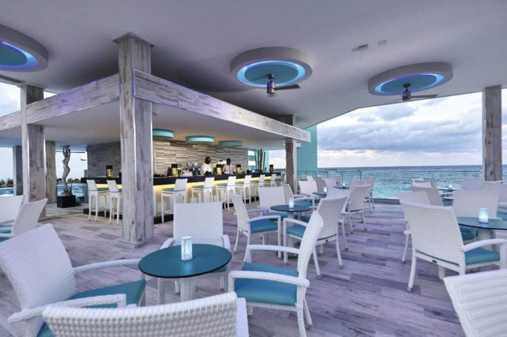 Bar ao ar livre e área de estar do Riu Palace Paradise Island com cadeiras de vime brancas e mesas azuis com vista para o oceano sob um céu limpo. Esse é um dos melhores resorts all inclusive no Caribe.