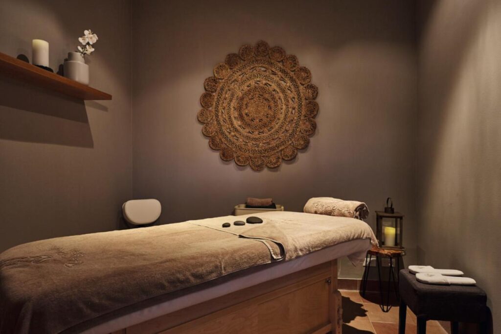 Uma sala de massagem no Riu Palace Paradise Island com uma mesa de tratamento, pedras, um suporte decorativo de parede, velas e toalhas em uma prateleira e no chão.