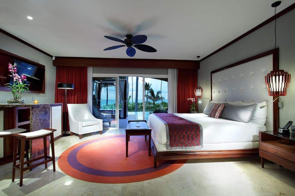 Um quarto moderno do Grand Palladium Bavaro Suites Resort & Spa com uma cama grande, área de estar e decoração em vermelho e branco. Uma porta de correr abre para uma vista do jardim tropical.