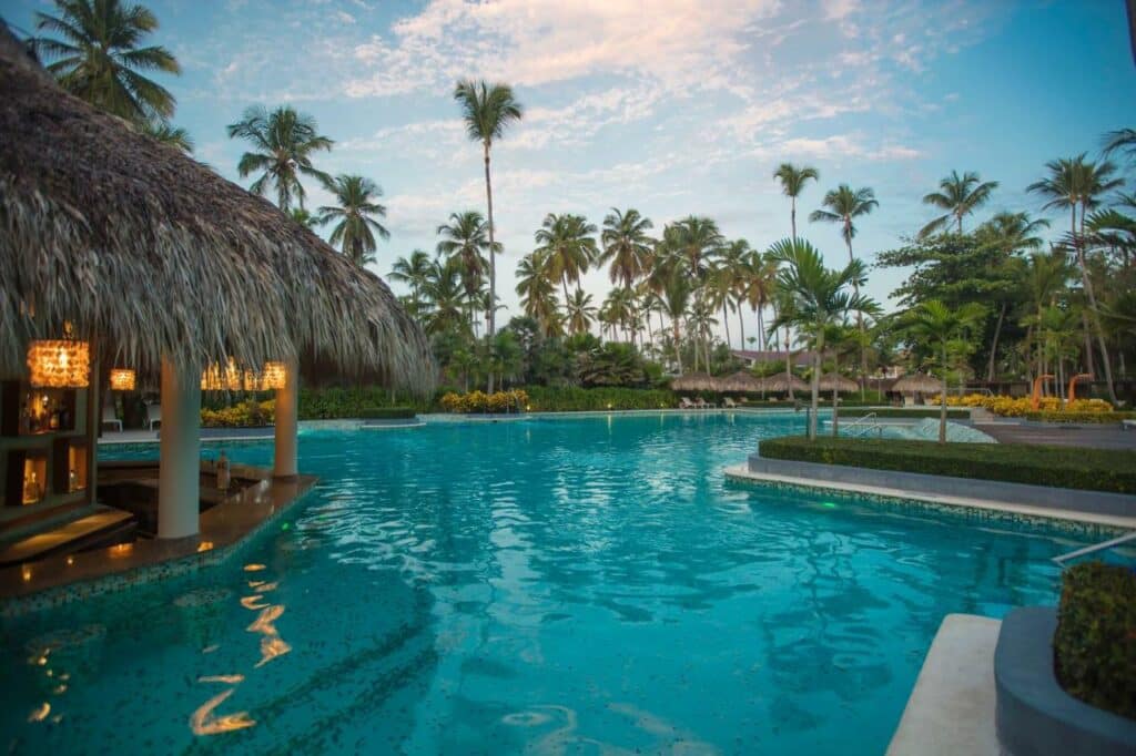 Uma piscina do Grand Palladium Bavaro Suites Resort & Spa com águas azuis cristalinas, cercada por palmeiras e estruturas com telhado de palha sob um céu parcialmente nublado. Esse é um dos melhores resorts all inclusive no Caribe.