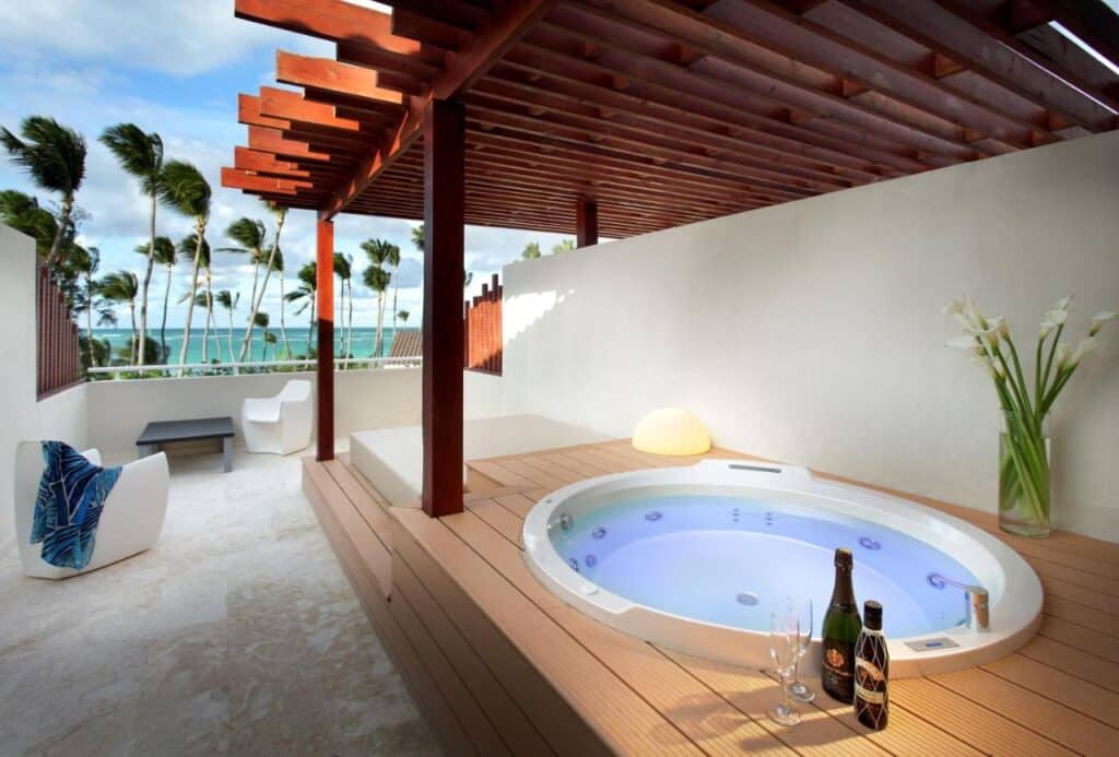 Terraço ao ar livre do Grand Palladium Bavaro Suites Resort & Spa com banheira de hidromassagem redonda, pérgola de madeira, cadeiras de praia e vista para o mar; garrafa de champanhe e taças por perto.