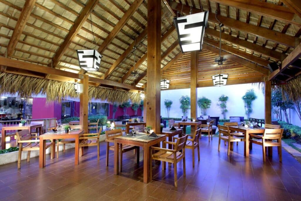 Restaurante ao ar livre do Grand Palladium Bavaro Suites Resort & Spa com mesas e cadeiras de madeira sob um teto de palha. Luzes suspensas modernas iluminam o espaço. Esse é um dos melhores resorts all inclusive no Caribe.