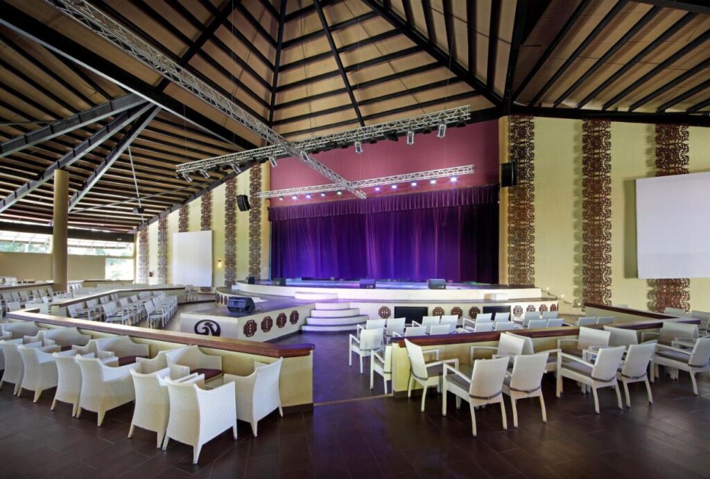 Um auditório do Grand Palladium Bavaro Suites Resort & Spa com assentos em camadas, cadeiras brancas, um palco com cortinas roxas e um teto alto de madeira.