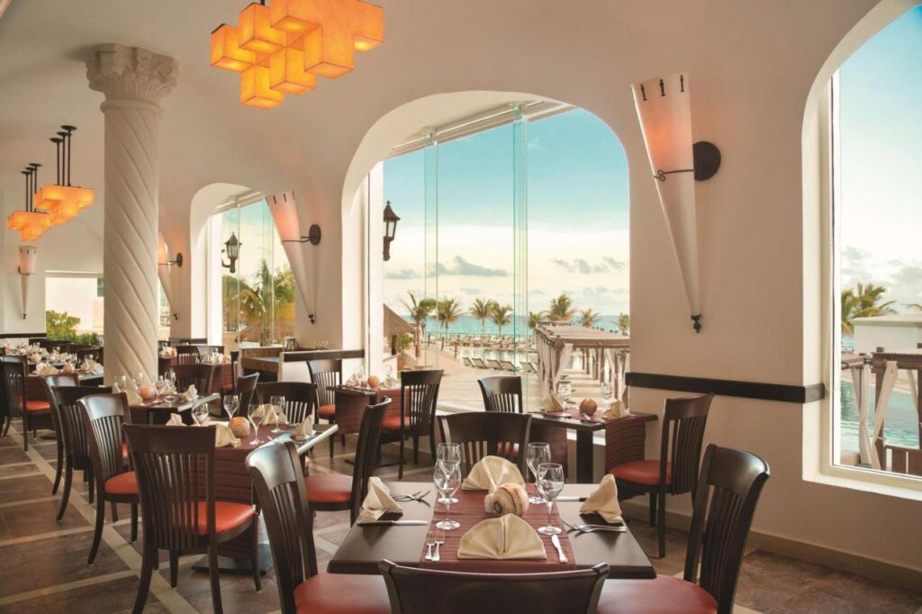 Restaurante elegante à beira-mar no Hyatt Zilara Cancun com mesas bem arrumadas, iluminação moderna e grandes janelas com vista para o oceano e as palmeiras.