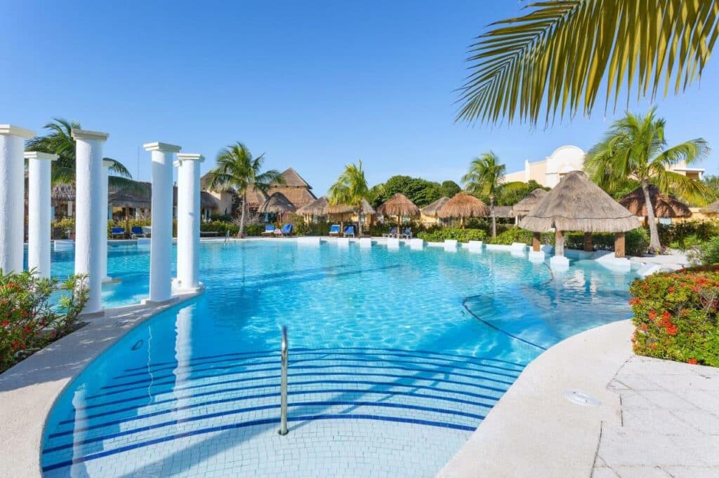 Uma piscina do Grand Palladium White Sand Resort & Spa com águas azuis cristalinas, palmeiras, cabanas de palha e colunas brancas sob um céu limpo. Esse é um dos melhores resorts all inclusive no Caribe.