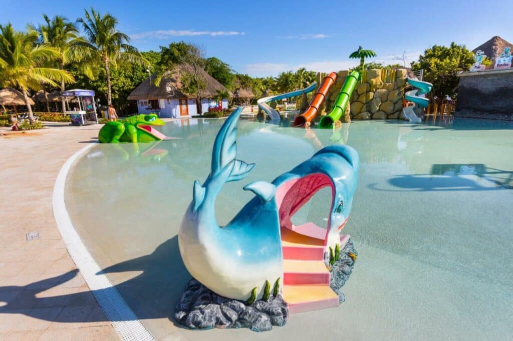 Um parque aquático infantil com toboáguas, um túnel de baleias e uma escultura de sapo cercada por palmeiras em um dia ensolarado no Grand Palladium White Sand Resort & Spa.