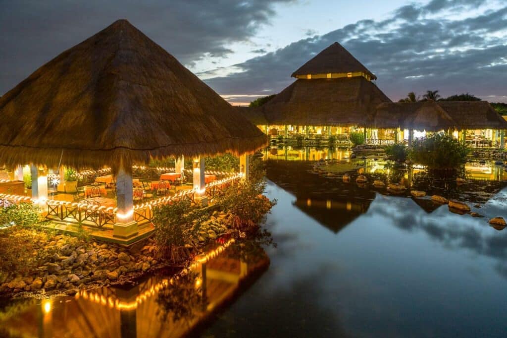 Edifícios com telhados de palha e luzes quentes são refletidos em um corpo de água calmo ao anoitecer, sob um céu nublado no Grand Palladium White Sand Resort & Spa.