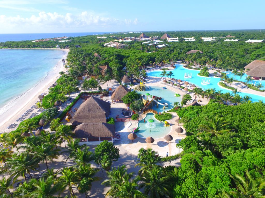 Vista aérea do Grand Palladium Colonial Resort & Spa com piscinas, palmeiras e edifícios com telhados de palha, adjacente a uma praia de areia branca. Esse é um dos melhores resorts all inclusive no Caribe.