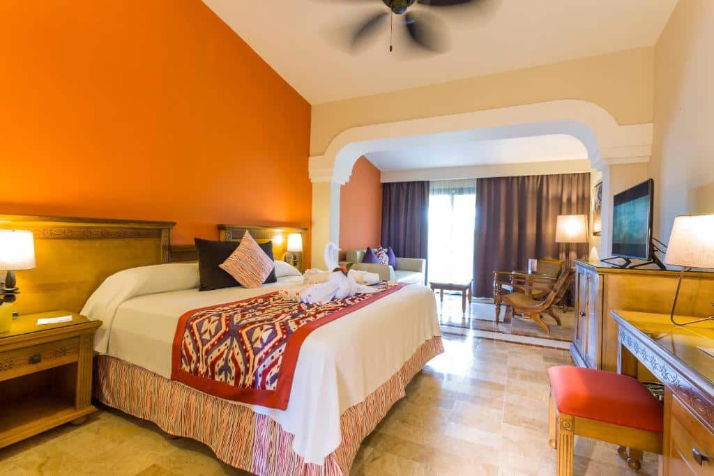 Um quarto do Grand Palladium Colonial Resort & Spa com uma cama grande, roupa de cama colorida, duas esculturas de cisnes em forma de toalha, uma área de estar, luminárias, uma TV e um ventilador de teto.