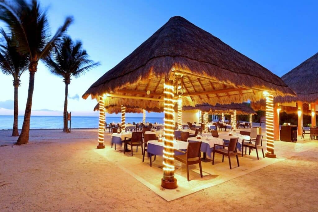 Restaurante à beira-mar no Grand Palladium Colonial Resort & Spa com telhado de palha e luzes de corda, cercado por palmeiras, com mesas dispostas para jantar à beira-mar ao pôr do sol. Esse é um dos melhores resorts all inclusive no Caribe.