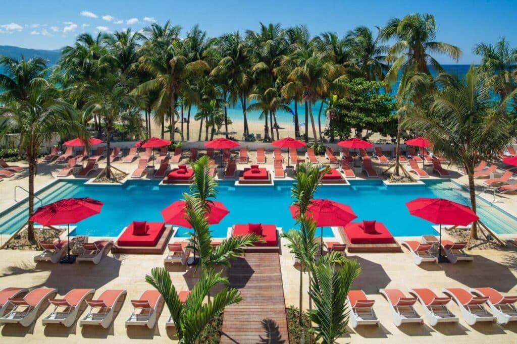 Uma área de piscina do S Hotel Montego Bay com espreguiçadeiras e guarda-sóis vermelhos, cercada por palmeiras, com vista para a praia e para o céu azul claro. Esse é um dos melhores resorts all inclusive no Caribe.