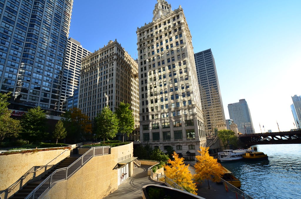 Prédios altos se alinham à margem de um rio em uma cidade, com uma ponte, barcos e algumas árvores amarelas de outono em primeiro plano. Imagem para ilustrar post sobre onde ficar em Chicago.