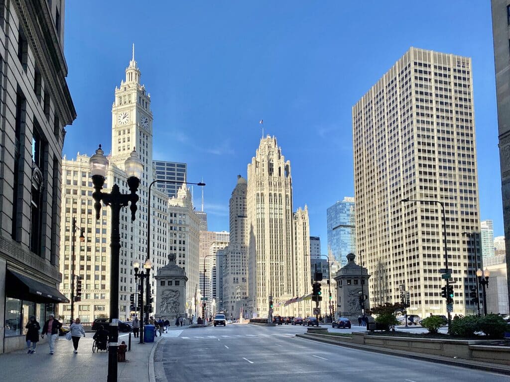 Rua no centro da cidade com altos edifícios históricos e modernos sob um céu azul, com tráfego leve e alguns pedestres. Imagem para ilustrar post sobre onde ficar em Chicago.