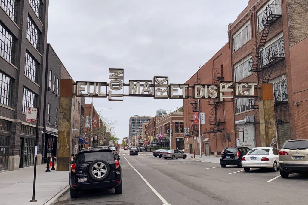Uma placa de metal com a inscrição "Fulton Market District" se estende por uma rua urbana repleta de carros estacionados e prédios de tijolos.