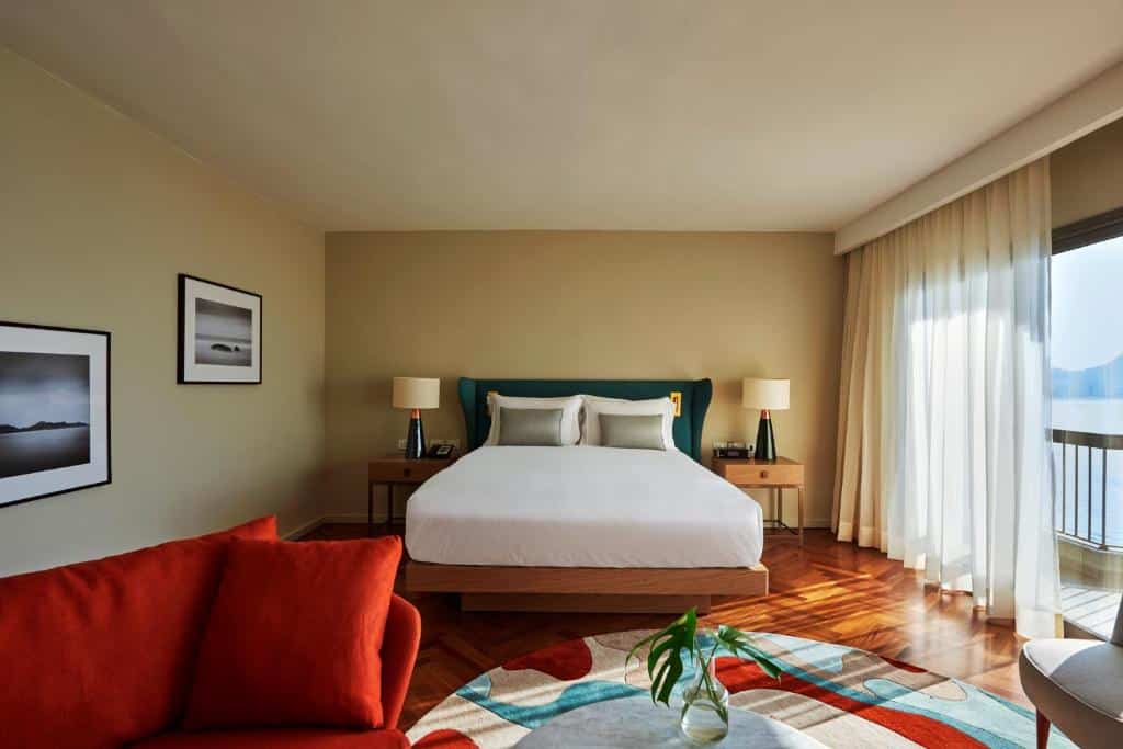 Um quarto moderno no Fairmont Rio de Janeiro Copacabana com uma cama grande, duas mesas de cabeceira, um sofá vermelho e janelas do chão ao teto com cortinas bege, para representar onde ficar em Copacabana.