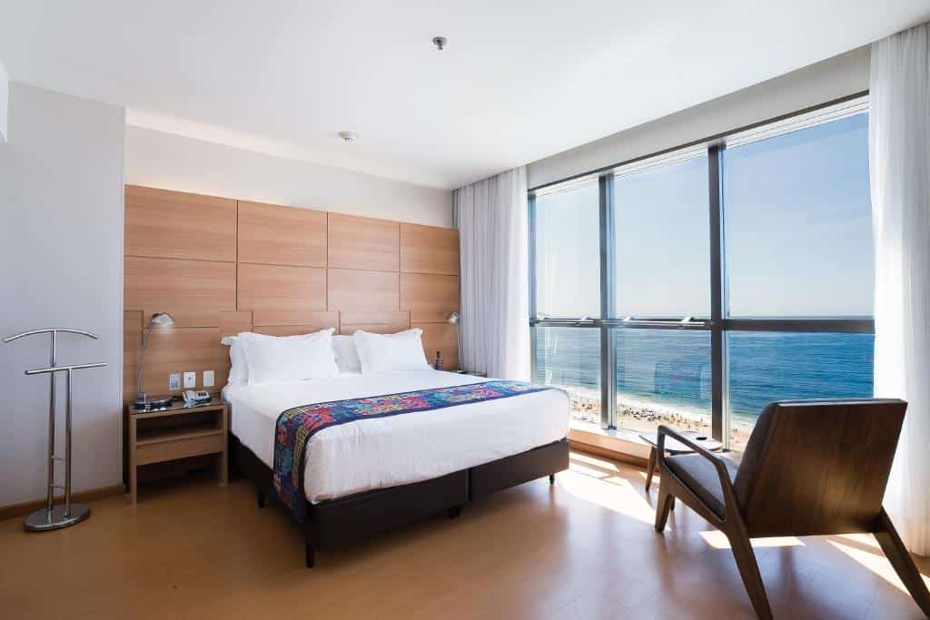 Quarto moderno no Arena Copacabana Hotel com uma cama de casal, cadeira e uma grande janela com vista para o oceano em um dia ensolarado, para representar onde ficar em Copacabana.