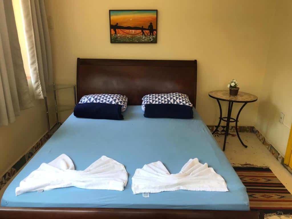 Uma cama bem arrumada com lençóis azuis, dois travesseiros e duas toalhas dobradas no pé, ao lado de uma mesa redonda e arte na parede, no Hostel Maresias do Leme, para representar onde ficar em Copacabana.