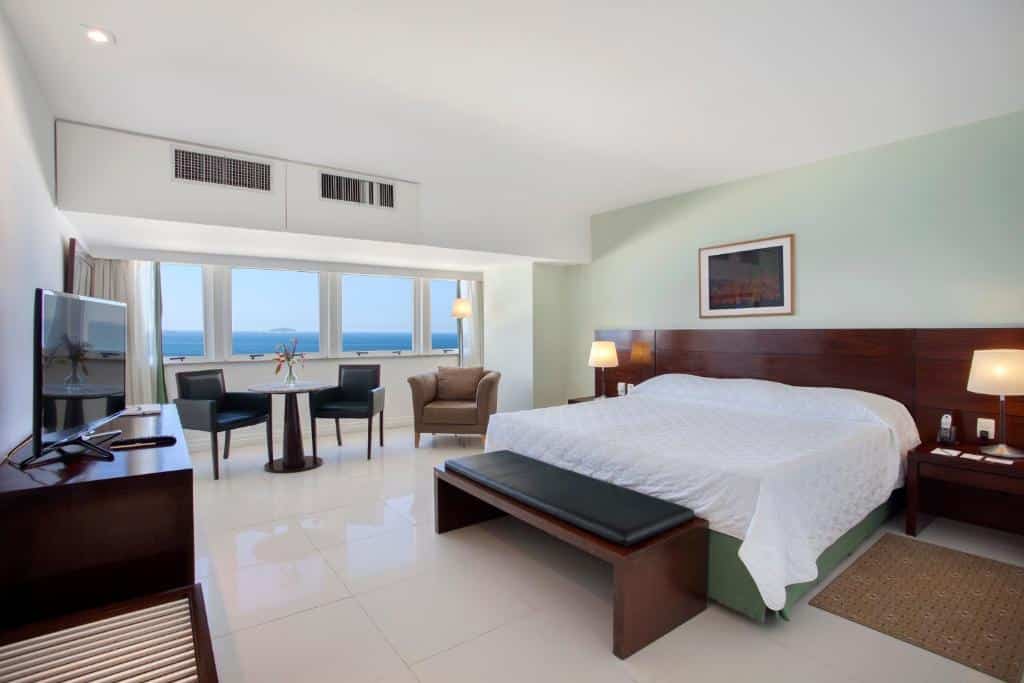 Quarto no Windsor Leme com uma cama grande, TV, área de estar e vista para o mar através de várias janelas, para representar onde ficar em Copacabana.