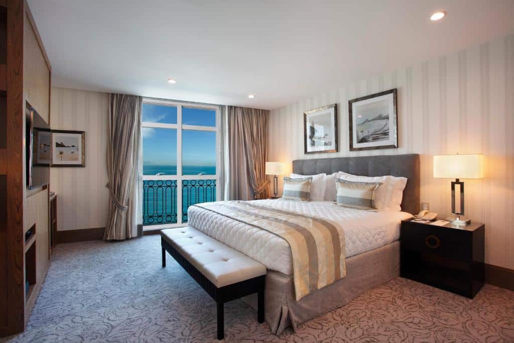 Quarto no Miramar Hotel by Windsor com uma cama grande, duas luminárias de cabeceira, um banco e uma janela com vista para o mar, para representar onde ficar em Copacabana.
