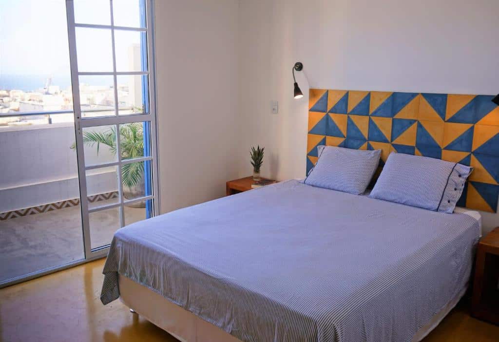 Um quarto no Aquarela do Leme com uma cama de casal, cabeceira geométrica, mesas de cabeceira e uma porta de vidro que leva a uma varanda com vista, para representar onde ficar em Copacabana.