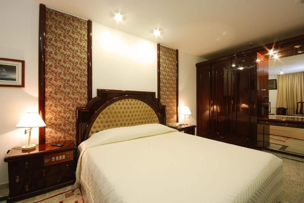 Uma cama bem arrumada com cabeceira tufada, mesas de cabeceira com luminárias e um grande guarda-roupa de madeira escura em um quarto do Américas Benidorm Hotel, para representar onde ficar em Copacabana.