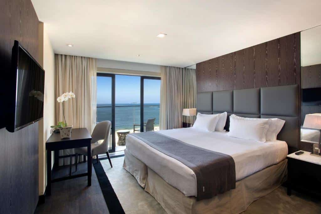 Quarto moderno no Windsor California Hotel com uma cama grande, escrivaninha, TV e uma varanda com vista para o oceano sob um céu claro, para representar onde ficar em Copacabana.