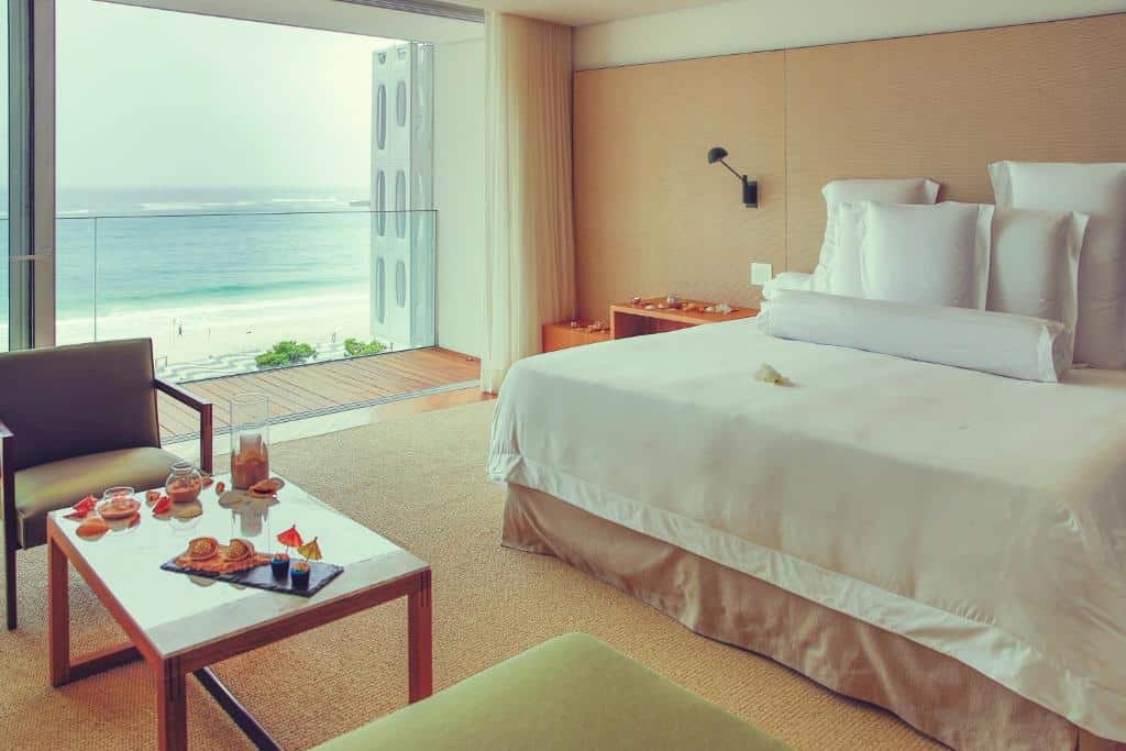 Quarto do Emiliano Rio com cama feita, poltrona, mesa com comida e varanda com vista para o mar, para representar onde ficar em Copacabana.