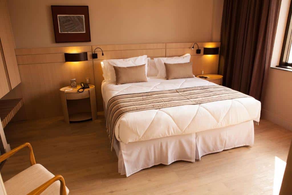 Uma cama de casal bem arrumada em um quarto no Ritz Copacabana Boutique Hotel com duas mesas de cabeceira, luminárias e uma janela com cortinas, para representar onde ficar em Copacabana.