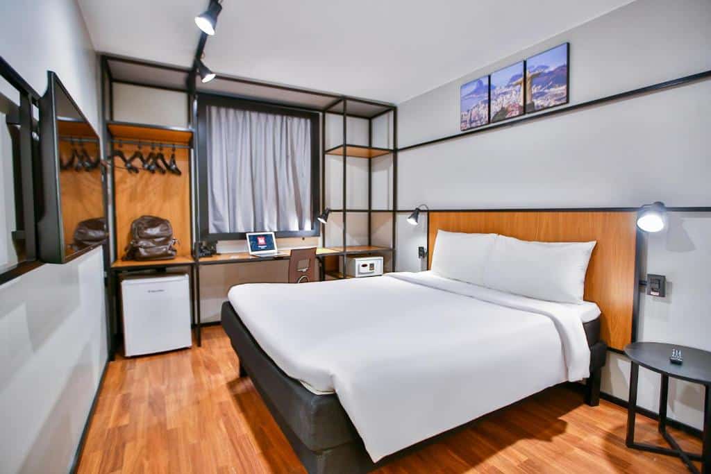 Quarto moderno no ibis Copacabana Posto 2 com cama de casal, escrivaninha, guarda-roupa aberto, frigobar e TV de parede com piso de madeira, para representar onde ficar em Copacabana.