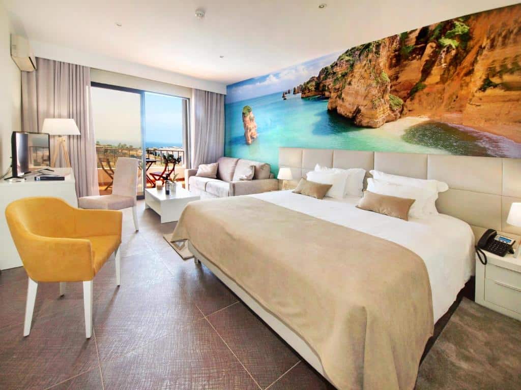 Quarto de hotel moderno com uma cama de casal, sofá, escrivaninha e um grande mural de parede de uma praia e falésias, além de uma varanda com vista.
