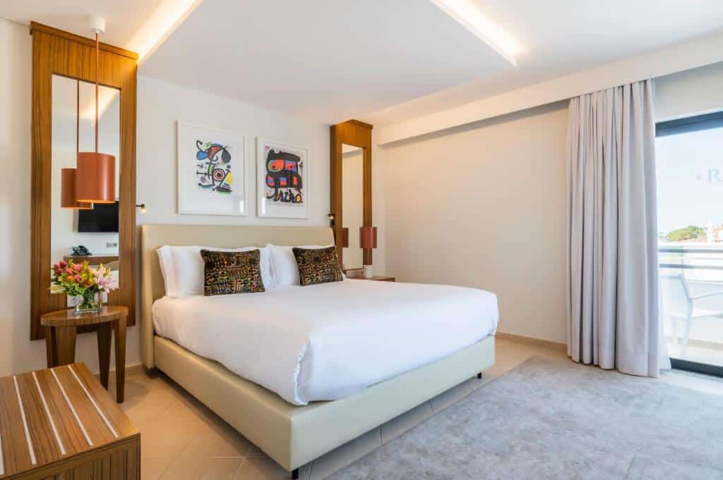 Quarto de hotel moderno com uma cama grande, mesas de cabeceira, arte na parede, flores e uma varanda com cortinas fechadas.