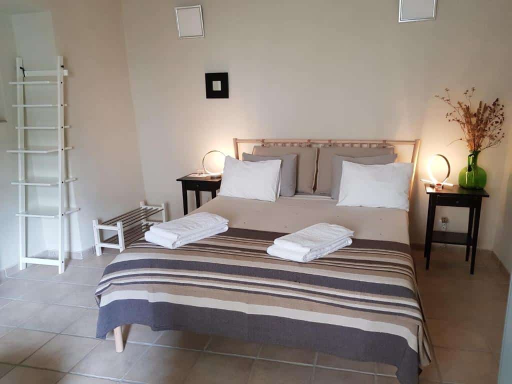 Uma cama de casal bem arrumada com roupa de cama listrada, duas toalhas, mesas de cabeceira e uma prateleira com escada em um quarto simples.