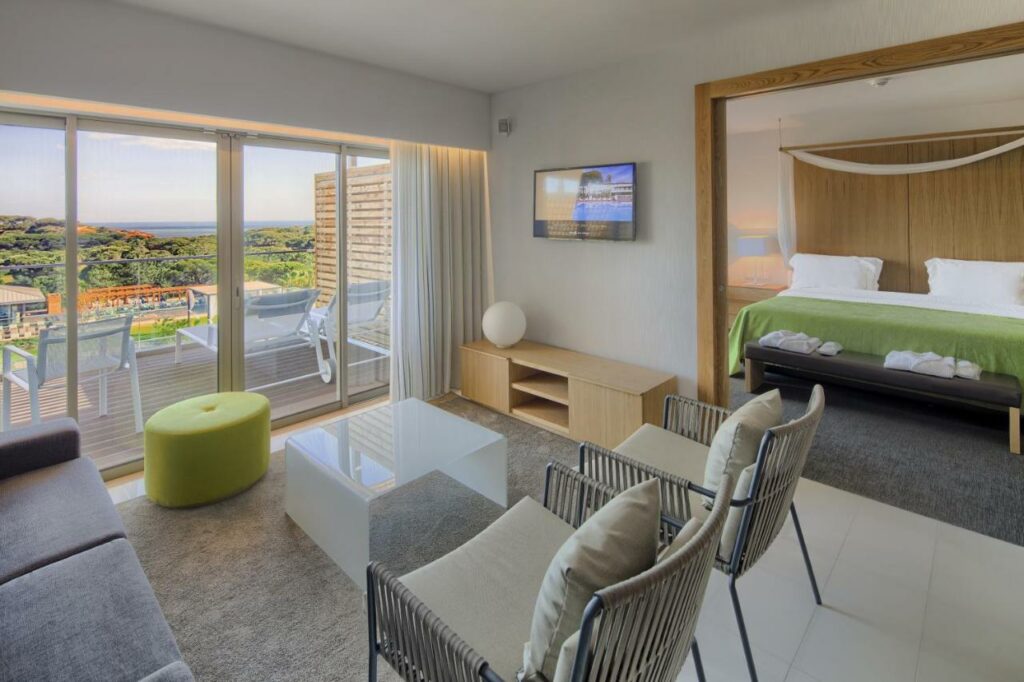 Suíte de hotel moderna com área de estar, varanda e quarto separado; as janelas grandes oferecem uma vista panorâmica do exterior. Esta é uma das indicações onde ficar no Algarve.