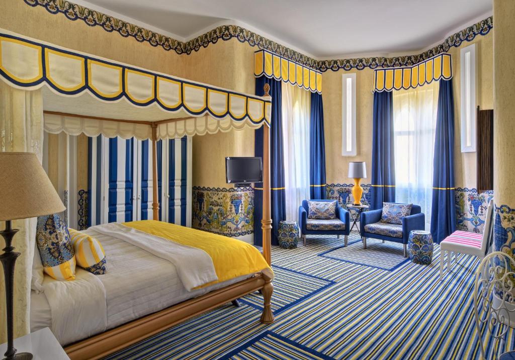 Quarto de hotel com decoração listrada em azul, amarelo e branco, cama de dossel, duas cadeiras e janelas grandes com cortinas.