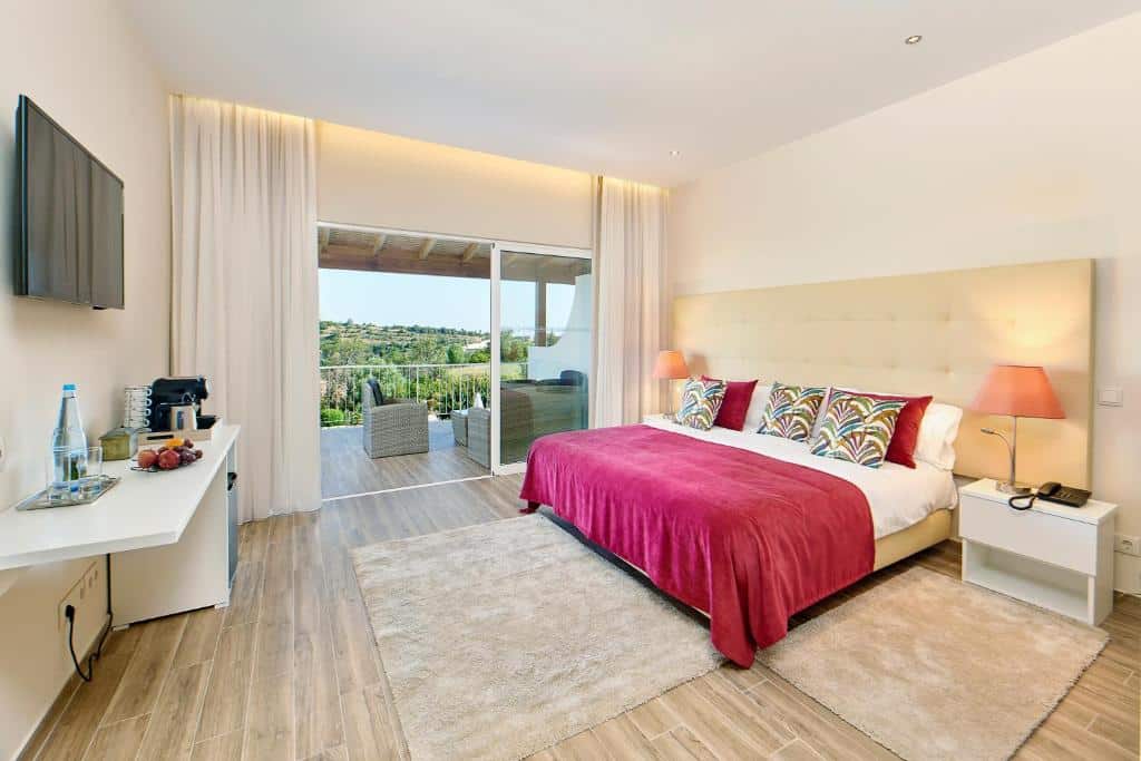 Quarto de hotel moderno com uma cama king-size, colcha vermelha, TV na parede, escrivaninha e varanda com assentos ao ar livre.