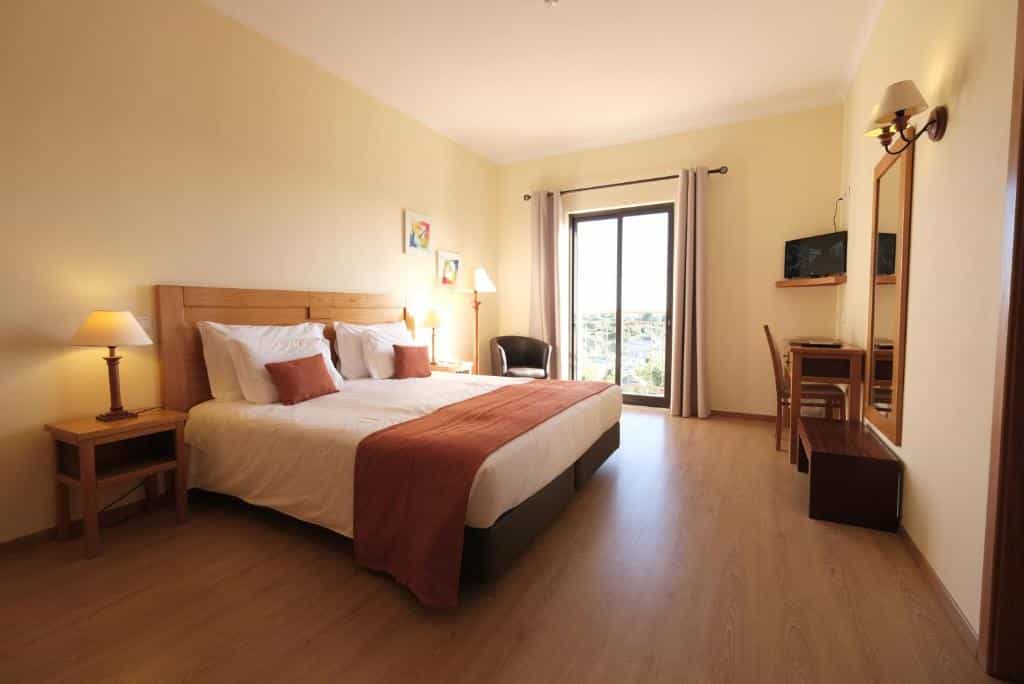 Quarto de hotel com uma cama de casal, duas mesinhas de cabeceira, uma escrivaninha, uma TV na parede e uma janela com cortinas.