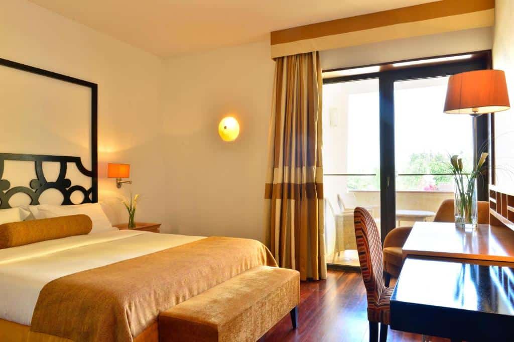 Um quarto de hotel com uma cama grande, escrivaninha, luminárias, uma cadeira e uma janela com cortinas que dão para uma varanda.