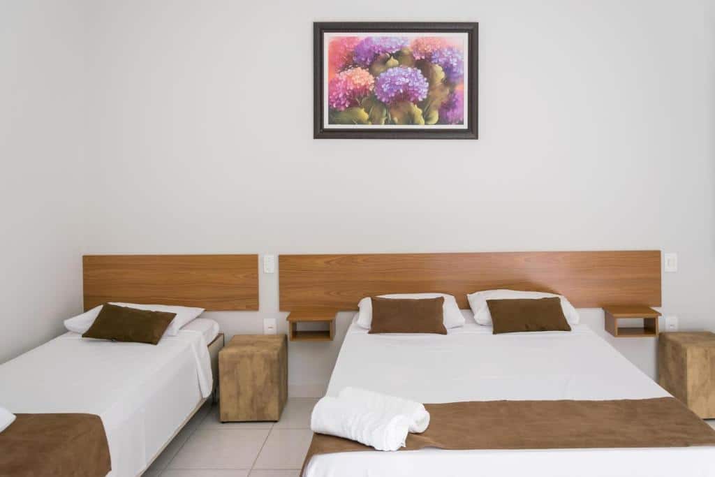 Um quarto na Pousada Flores do Campo com uma cama de casal e uma cama de solteiro, ambas com detalhes em marrom, e uma pintura floral acima das camas, para rperesentar pousadas em Águas de Lindóia.
