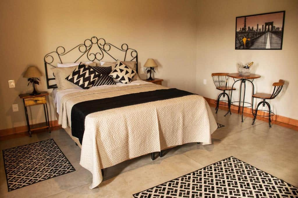 Quarto na Pousada Estância das Montanhas com cama de casal, travesseiros geométricos, mesinhas, duas cadeiras com uma mesa redonda e arte na parede, para representar pousadas em Águas de Lindóia.