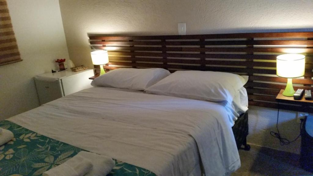 Uma cama bem arrumada com lençóis brancos, dois travesseiros e duas lâmpadas de cabeceira acesas em um quarto suavemente iluminado, na Vista da Serra Pousada de Águas.