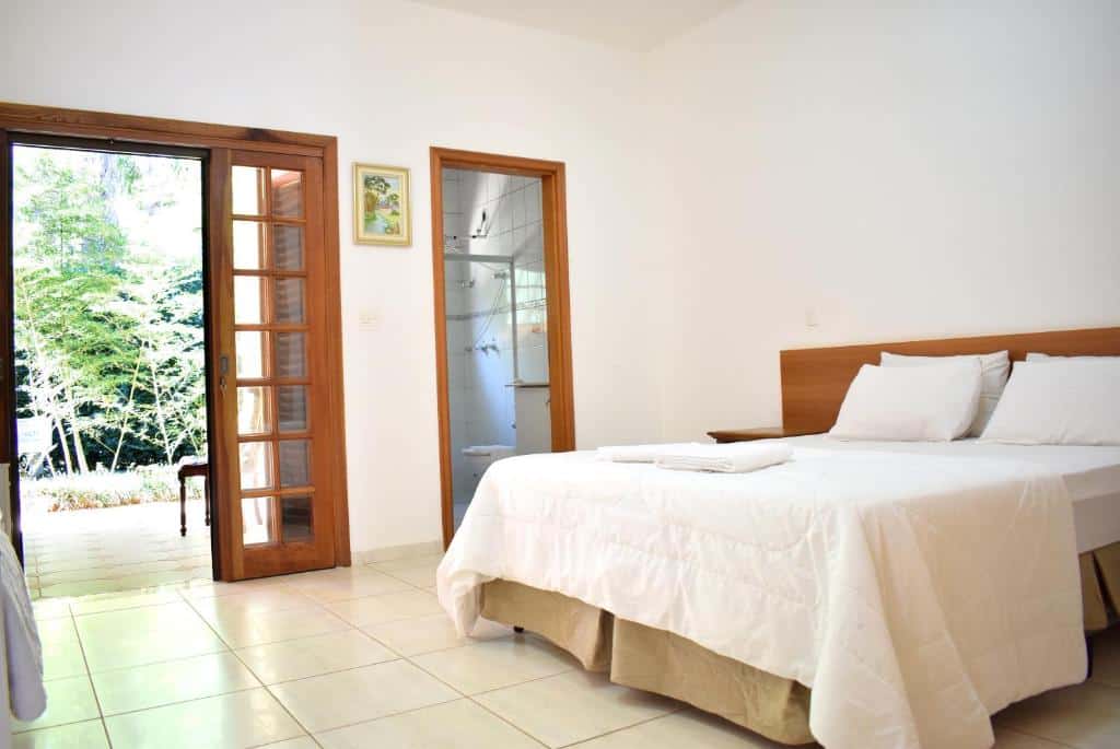 Um quarto arrumado na Pousada Appaloosa com uma cama de casal, roupa de cama branca, uma porta aberta para um pátio e uma vista para o banheiro, para representar pousadas em Águas de Lindóia.