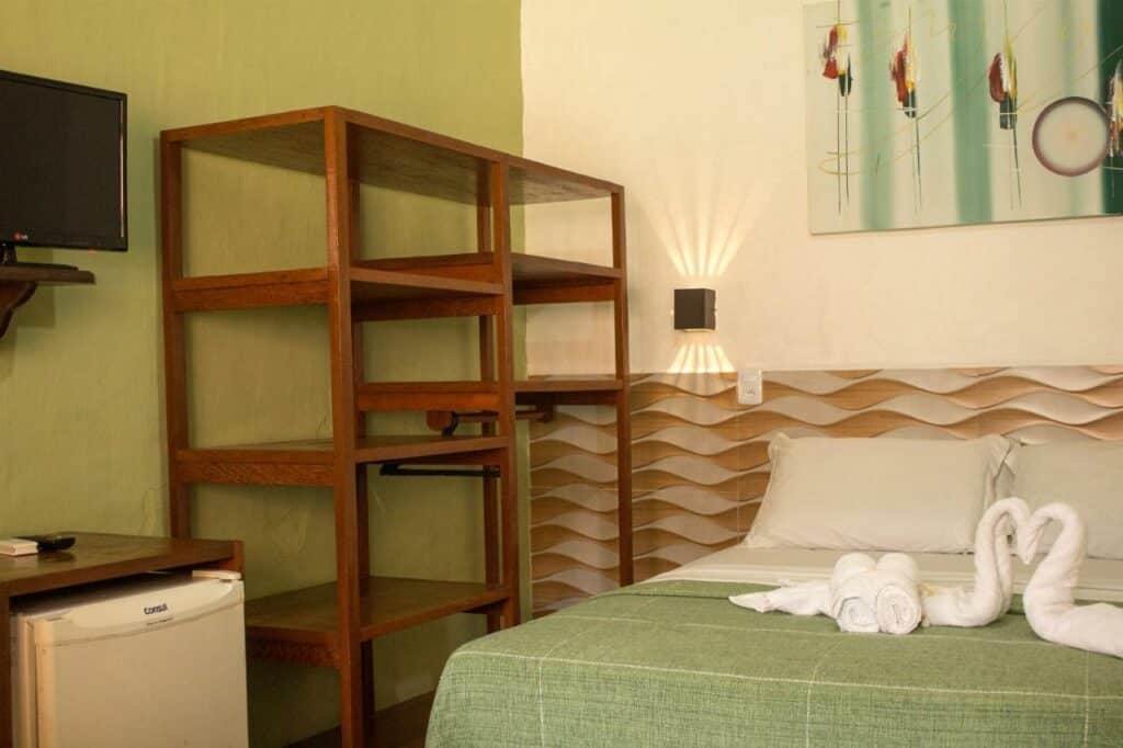 Quarto da Pousada Cavalo Marinho. No canto direito da foto há uma cama com lençol verde e travesseiros brancos. Toalhas dobradas em formato de coração estão em cima da cama, e há um quadro a parede acima, bem como um abajur ao lado.
Na parede esquerda, uma estante de madeira com divisórias está ao lado de um frigobar. Há uma televisão na parede acima.