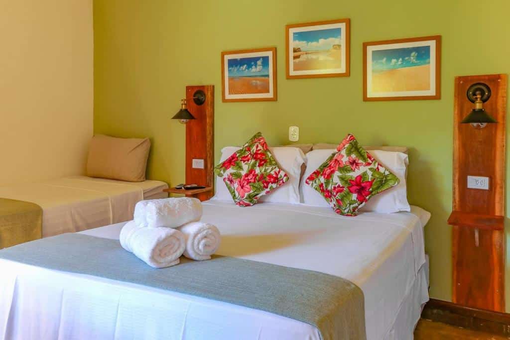 Quarto da Pousada Catamarã. Uma cama de casal tem lençol e travesseiros brancos, assim como algumas toalhas enroladas aos pés. Duas almofadas floridas estão na frente dos travesseiros. Há três quadrinhos na parede acima da cama, e luminárias acima do apoio de cabeceira dos dois lados.
Há uma cama de solteiro com lençol e travesseiro do lado esquerdo do quarto.