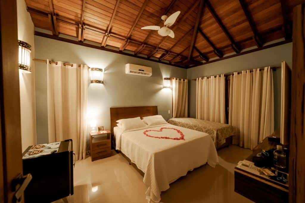 Quarto da Pousada Arraial Charme, uma das recomendações de pousada em Arraial D'Ajuda. Há uma cama de casal com lençol e travesseiros brancos no centro do quarto. Um coração de pétalas de rosa está no meio da cama. Há uma mesinha de cabeceira do lado esquerdo e uma cama de solteiro do lado direito.
Na parede esquerda tem um frigobar preto. Na frente da cama há uma mesa com uma televisão na parede acima.
Há um ventilador de teto no quarto, assim como um ar-condicionado e janelas com cortinas brancas que vão até o chão.