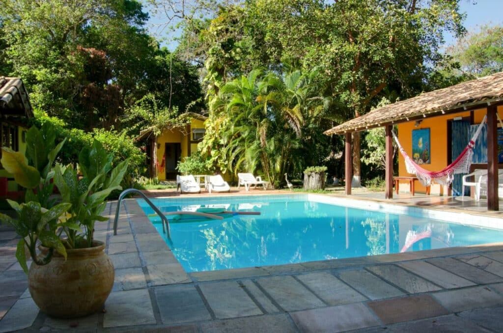 Piscina da Pousa Macunaíma, uma das recomendações de pousadas em Arraial D'Ajuda. Há diversas árvores e plantas ao redor, e espreguiçadeiras brancas se estendem pela lateral da piscina.
Do lado direito há uma área coberta com rede e cadeiras.